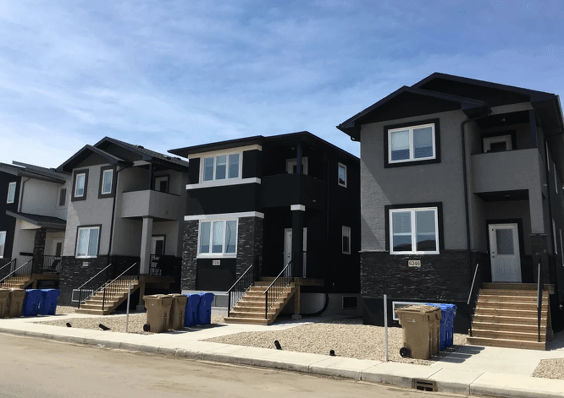A street of finished two-storey multi-unit homes in grey, black and stone cladding, each with its own front stairs and walkway, bins at the curb under a blue sky