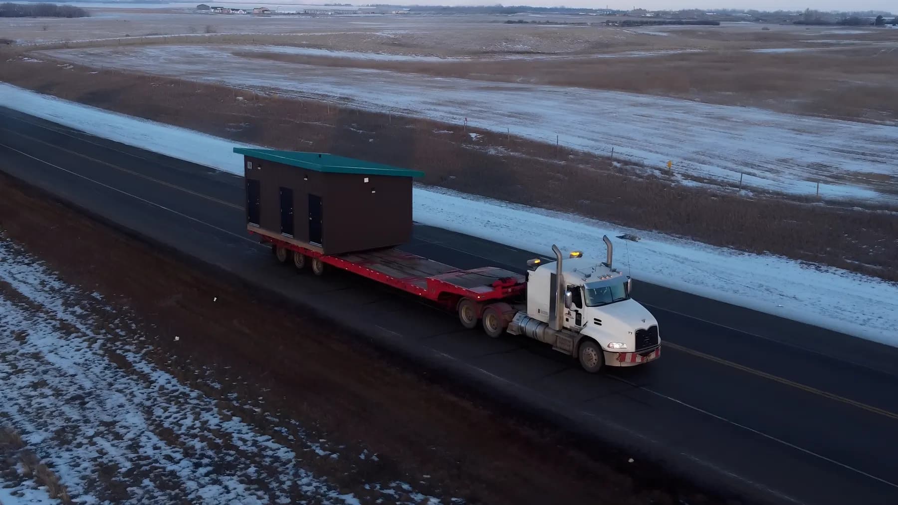 An aerial view of a semi hauling a dark brown modular building with a green roof and three doors along a two-lane highway through snow-dusted prairie
