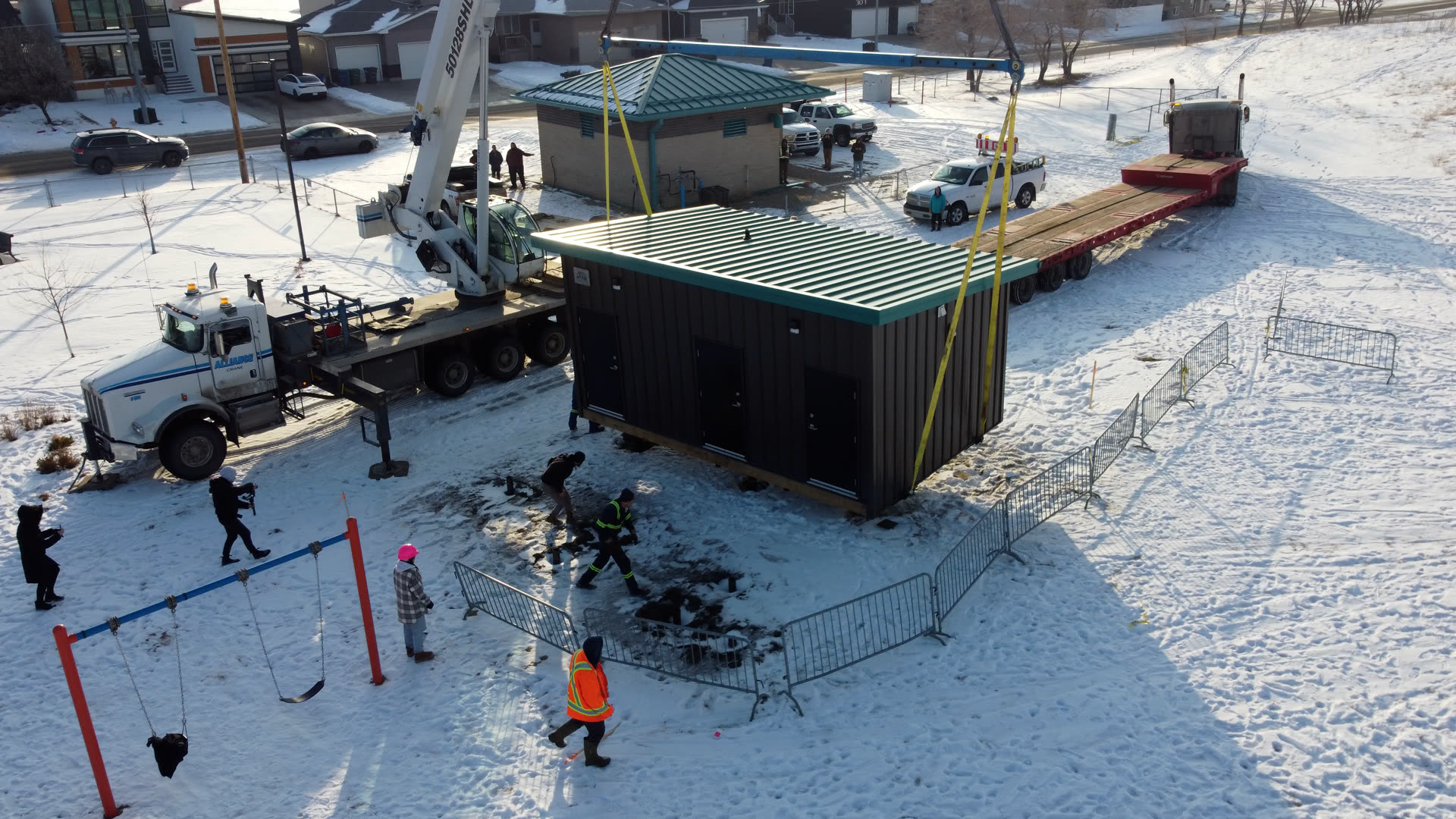 A crane sets a finished dark-clad Herauf modular unit onto its foundation at a prepared winter site