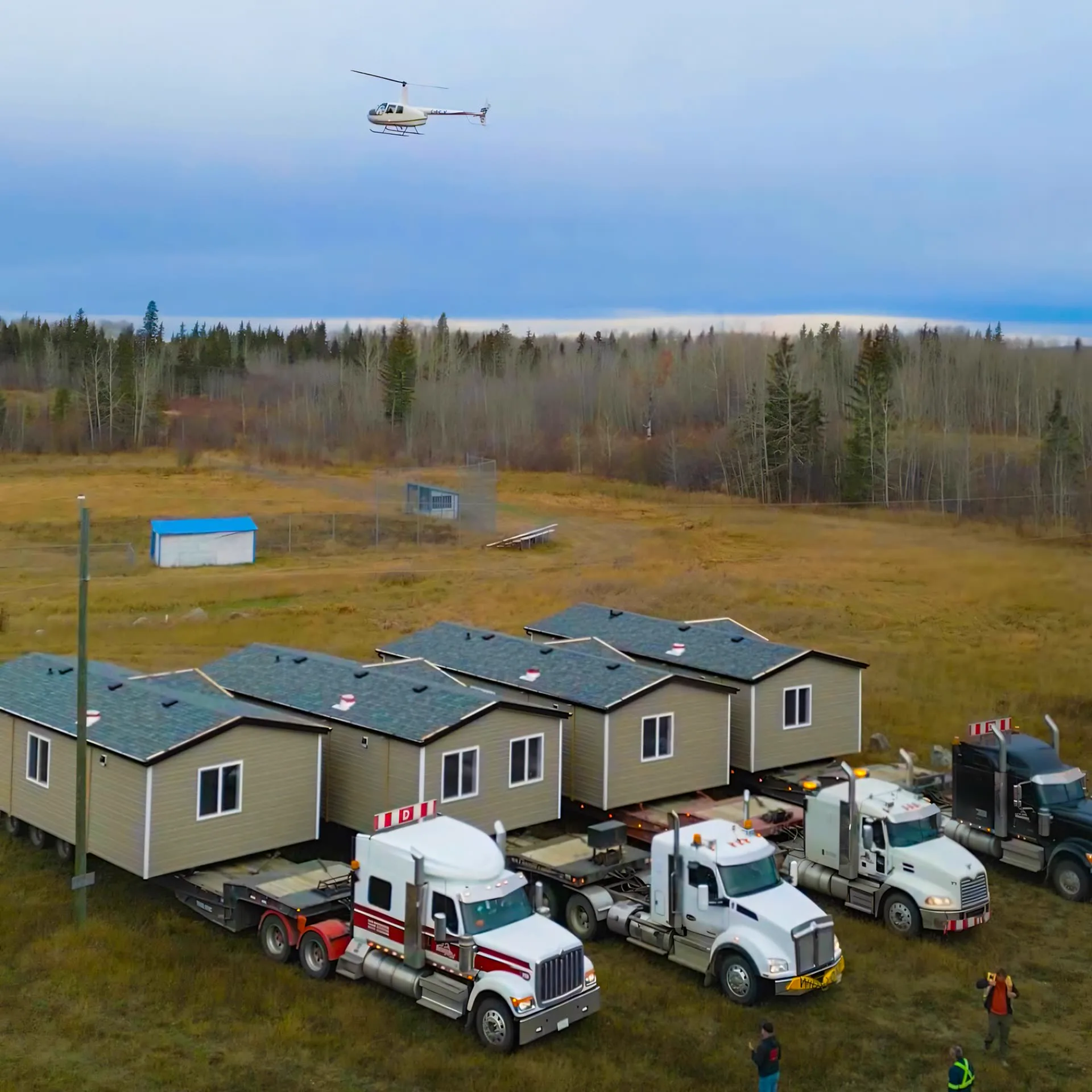 Four finished modular units with grey shingle roofs on lowboy trailers hitched to a row of semi trucks in a forest clearing, crew on the ground and a helicopter overhead