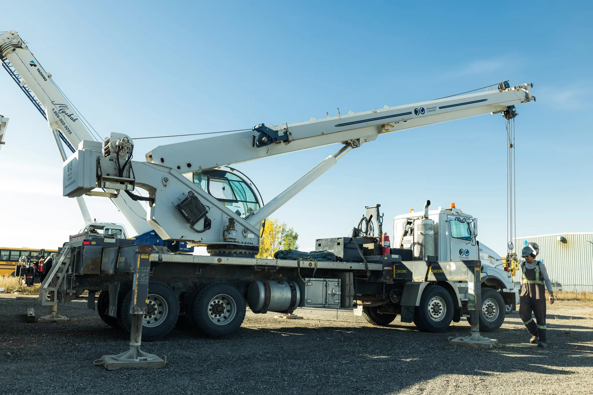 A boom truck crane with its outriggers down in the gravel yard, a crew member in high-visibility gear beside it