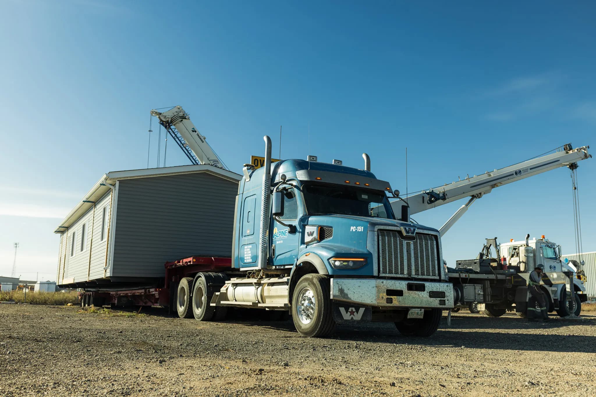 A blue highway tractor hitched to the loaded module, a crane on each side