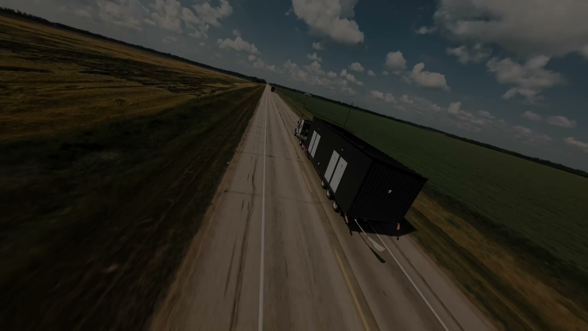 A low aerial chase of a semi hauling a dark-clad modular building down a two-lane prairie highway between green and harvested fields