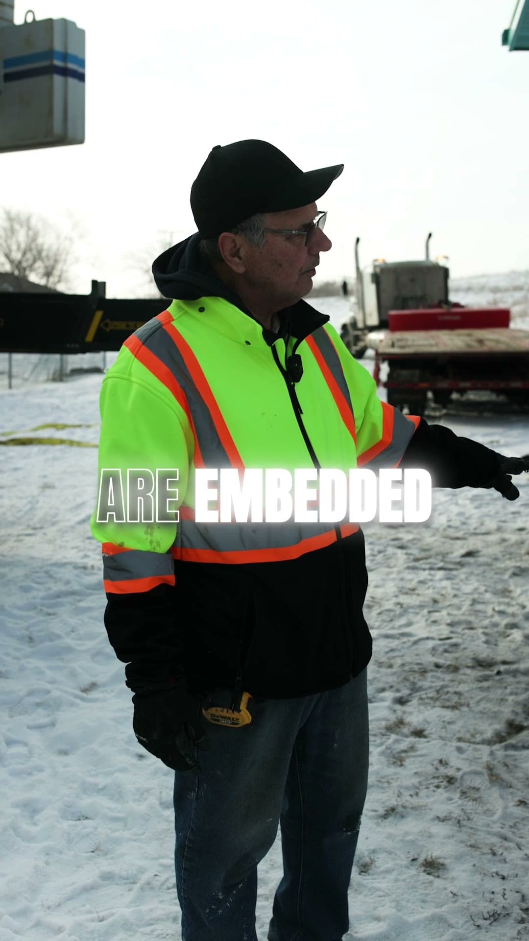 Crew member in a hi-vis jacket and cap talking on a snowy lot, a loaded truck behind him