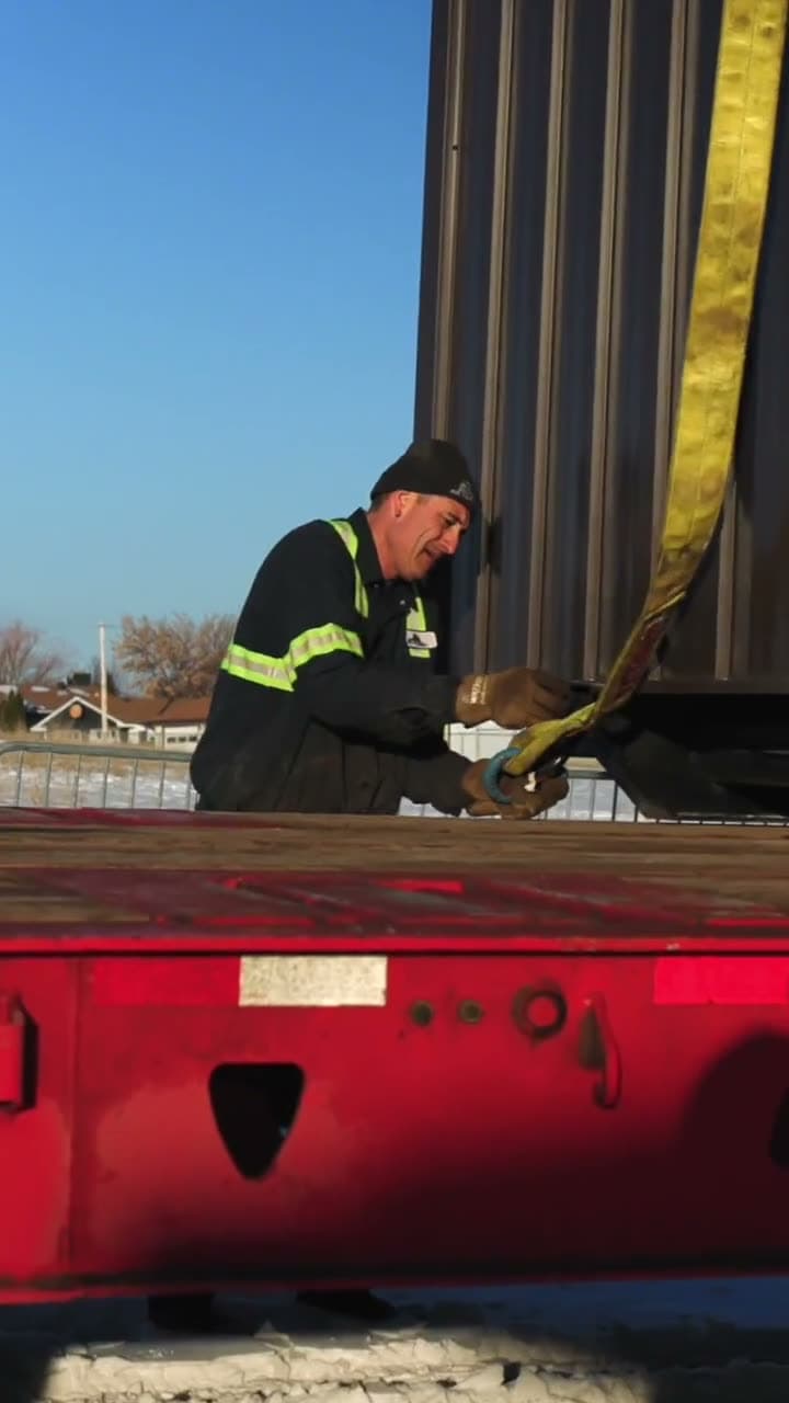 Crew member in a hi-vis jacket tightening a strap on a loaded flatbed trailer