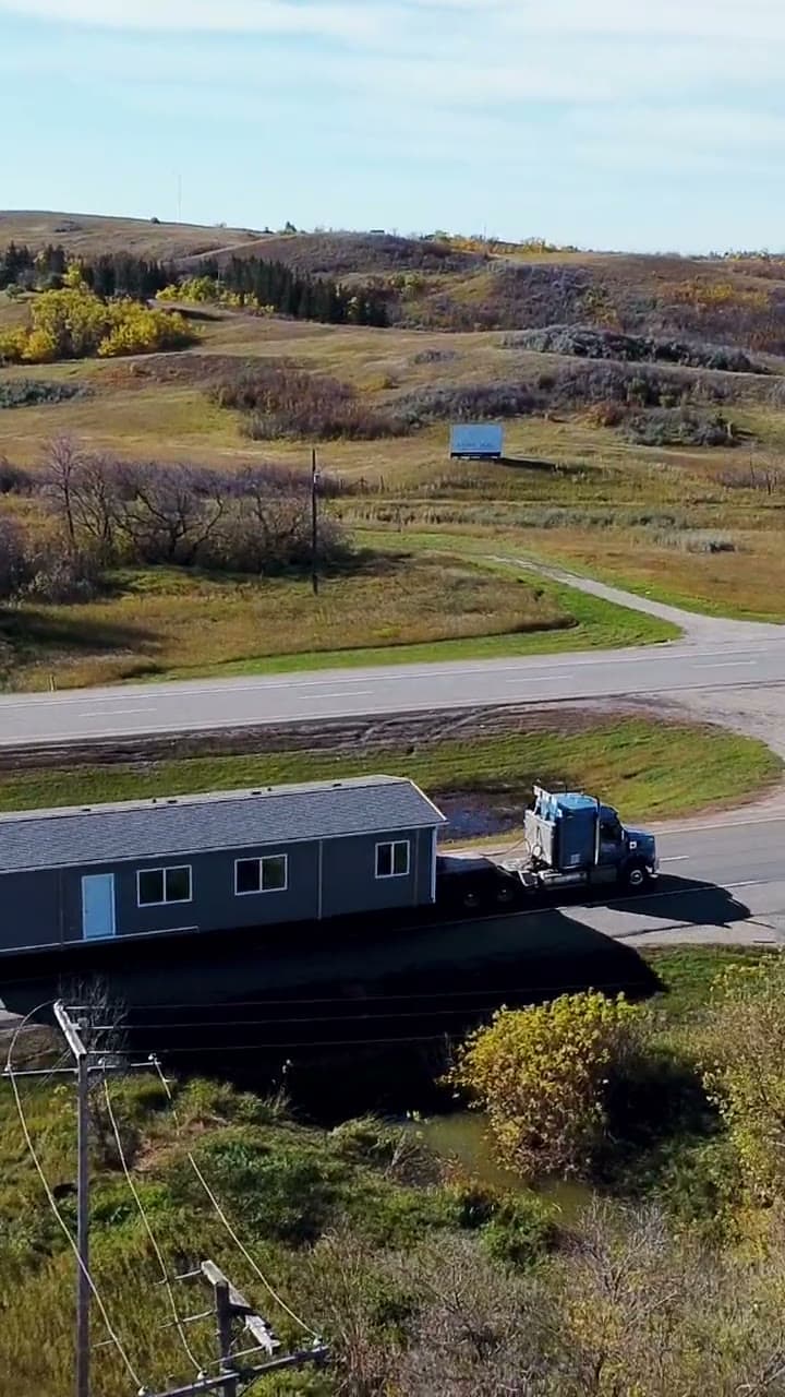 Drone view of a blue truck hauling a grey modular building along a highway past autumn hills