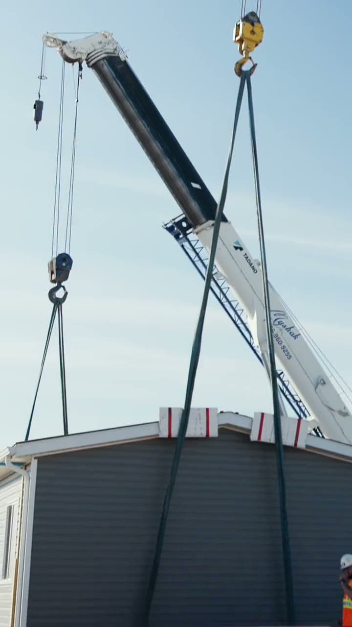 A crane hook and slings lifting a grey modular building off the ground, a second crane boom behind it
